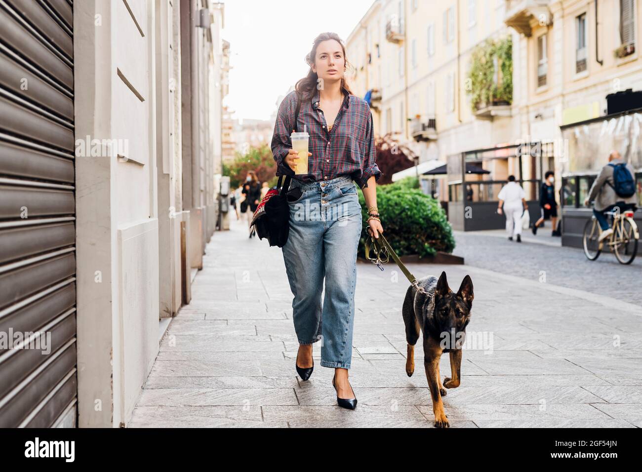 Woman holding drink while walking with German shepherd dog on footpath in city Stock Photo