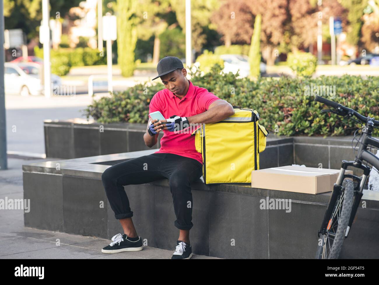 Delivery man text messaging through smart phone while sitting on bench by backpack Stock Photo