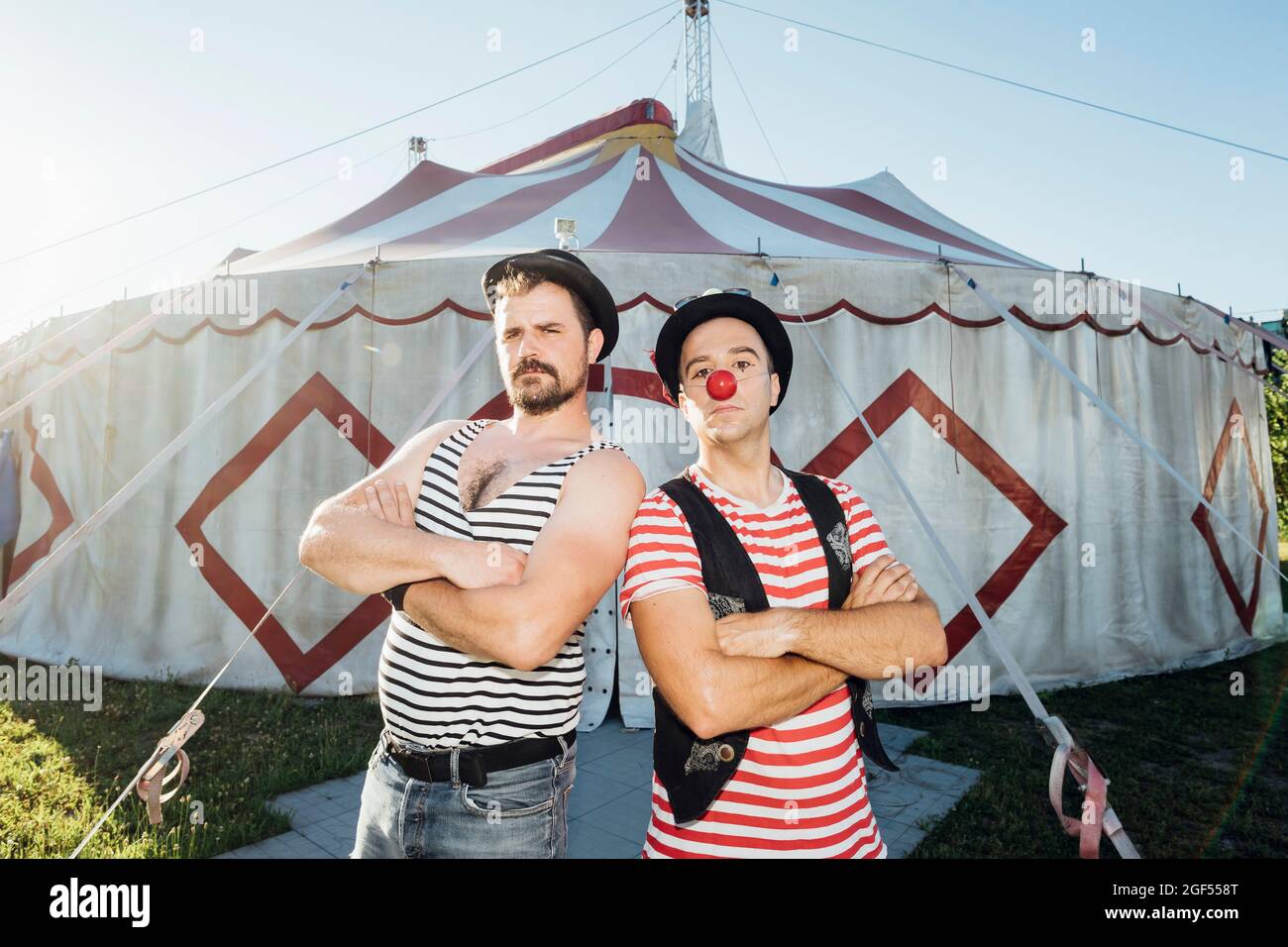 Muscular build artist standing with arms crossed by clown in front of circus tent Stock Photo