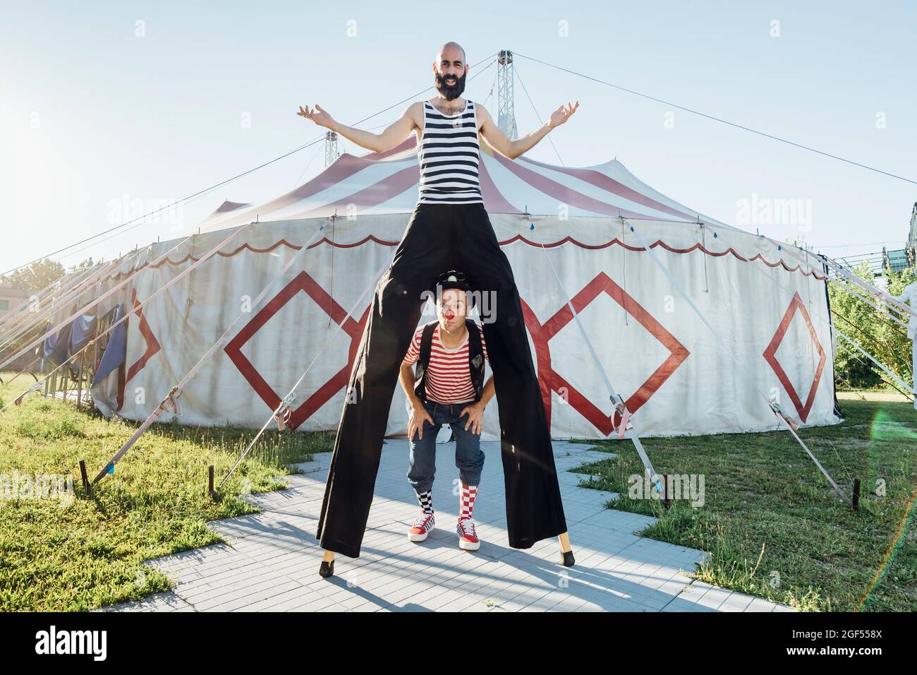 Male clown standing with performer on stilts in front of circus tent
