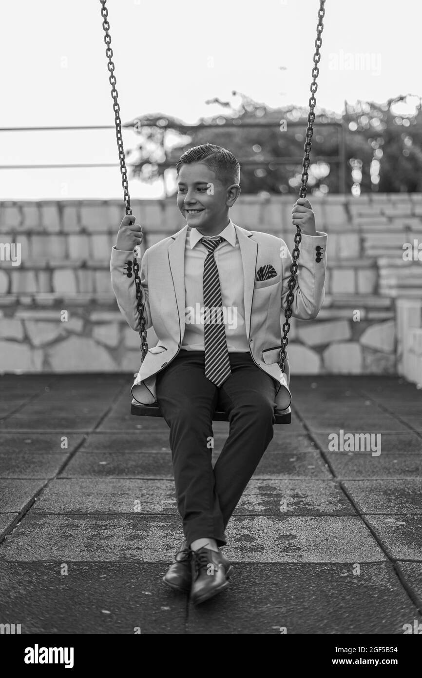 Vertical grayscale shot of an adorable male child in a formal suit outfit posing on a swing Stock Photo