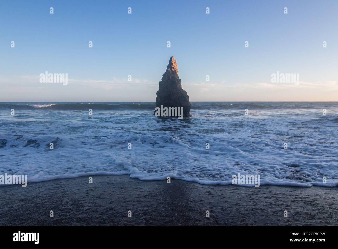 A wide angle landscape shot of the ocean and beach with a tall, pointy rock off the shore coast with waves rolling onto the shoreline at sunset Stock Photo