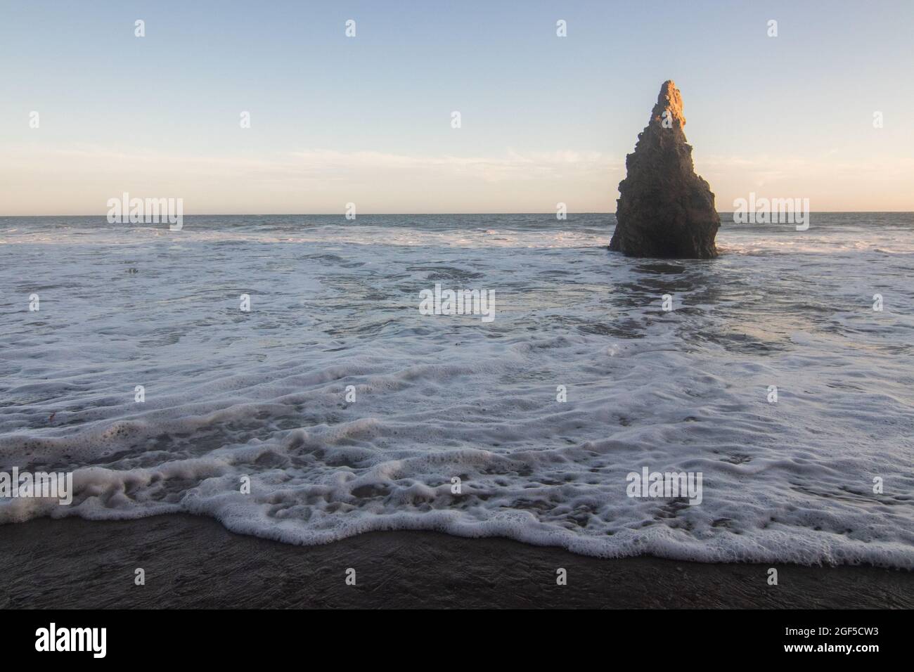 A wide angle landscape shot of the ocean and beach with a tall, pointy rock off the shore coast with waves rolling onto the shoreline at sunset Stock Photo