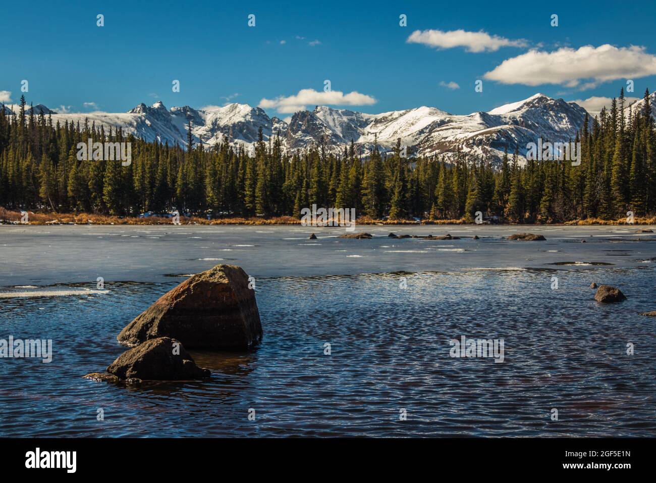 A wide angle landscape shot of a snowy Colorado mountain range and pine trees with a lake of water in the foreground with rocks, daytime in winter Stock Photo