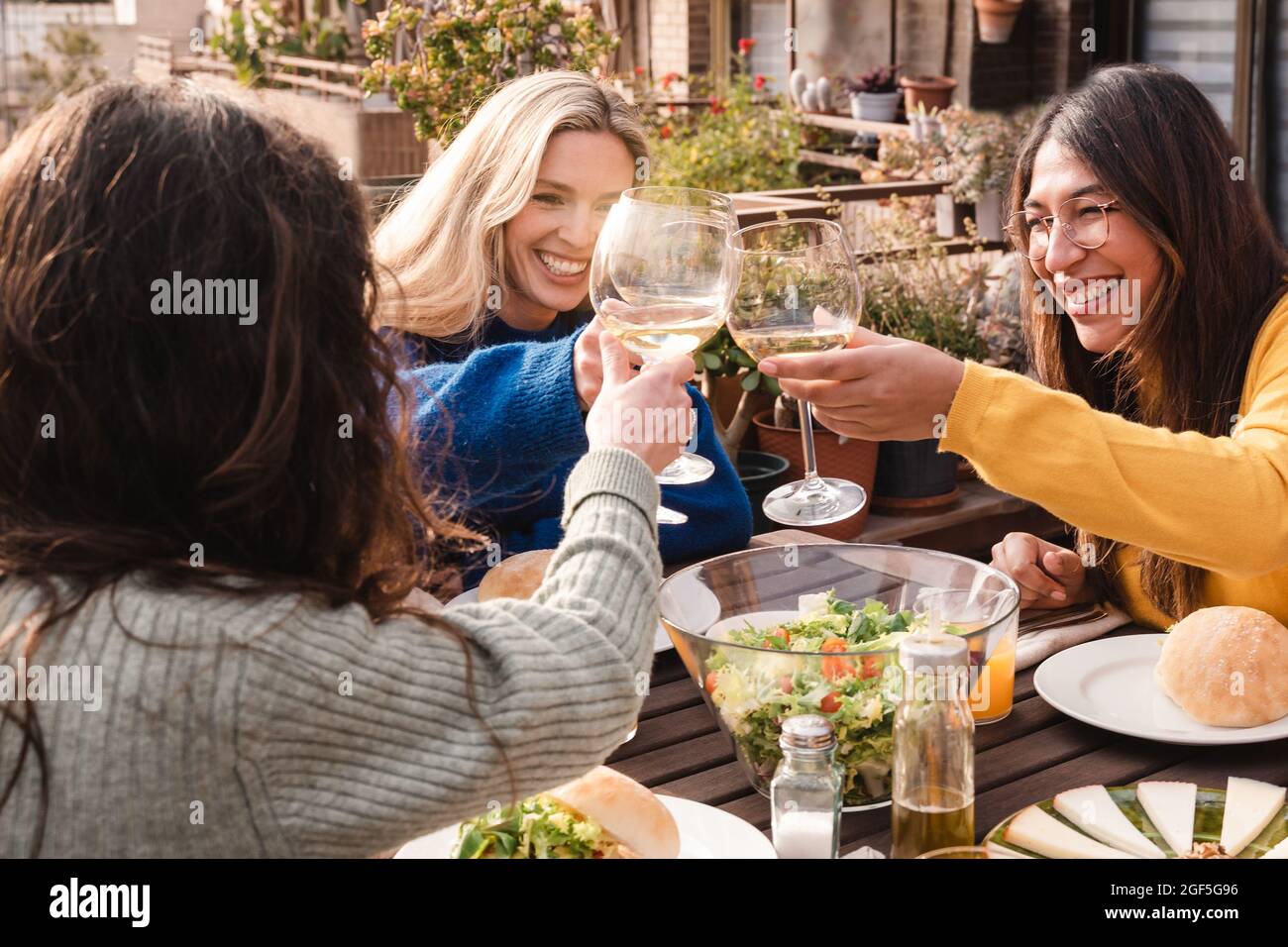 Multiracial friends eating vegan food cheering with wine outdoors at patio restaurant - Focus on center girl eye Stock Photo