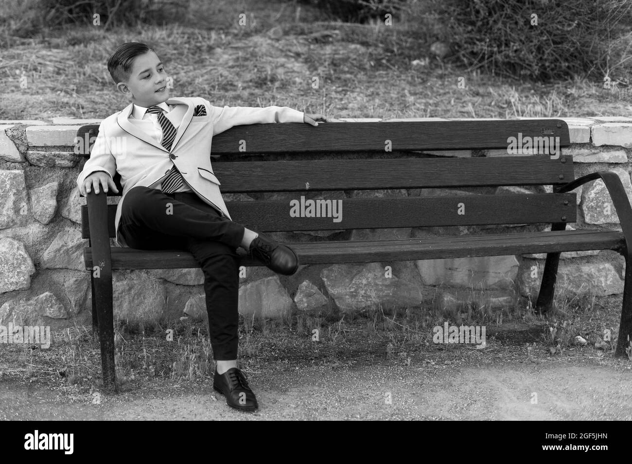 Grayscale shot of an adorable male child in a formal suit outfit posing on a bench Stock Photo