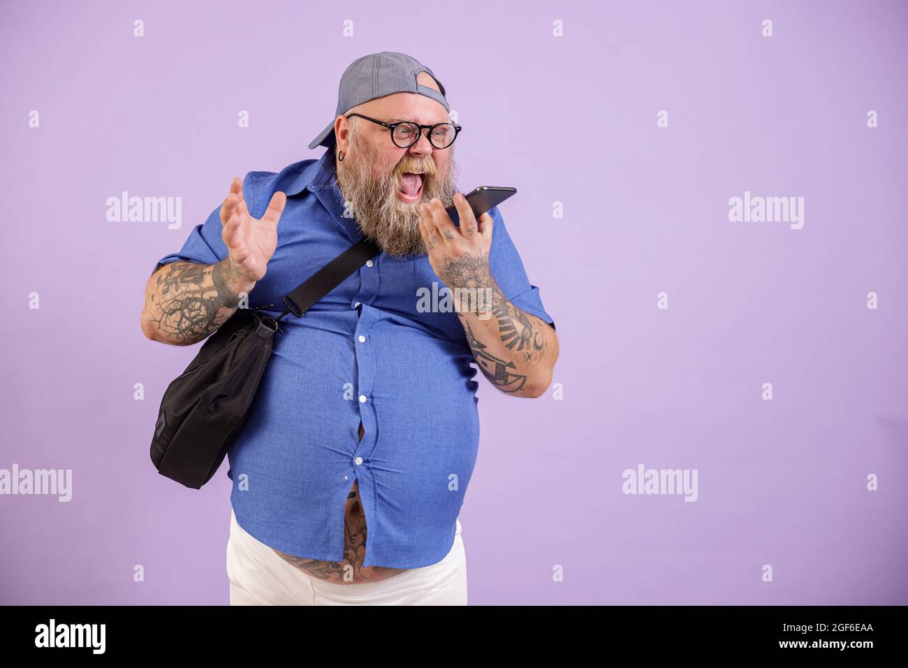 Angry man with overweight yells by phone using loudspeaker mode on purple background Stock Photo