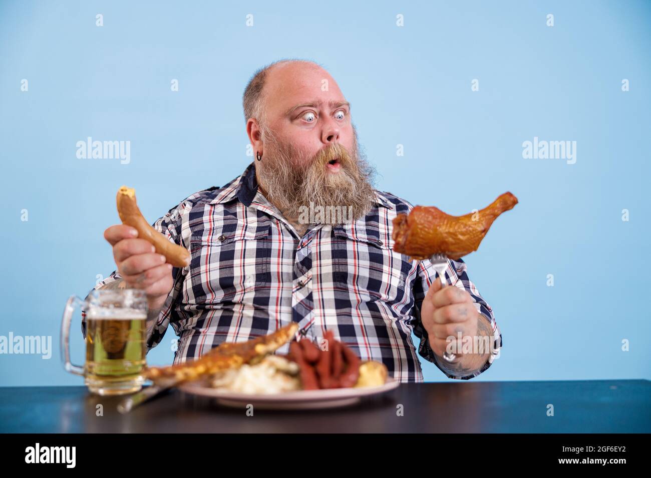 Shocked plus size man looks at chicken leg at table with fat food and beer Stock Photo