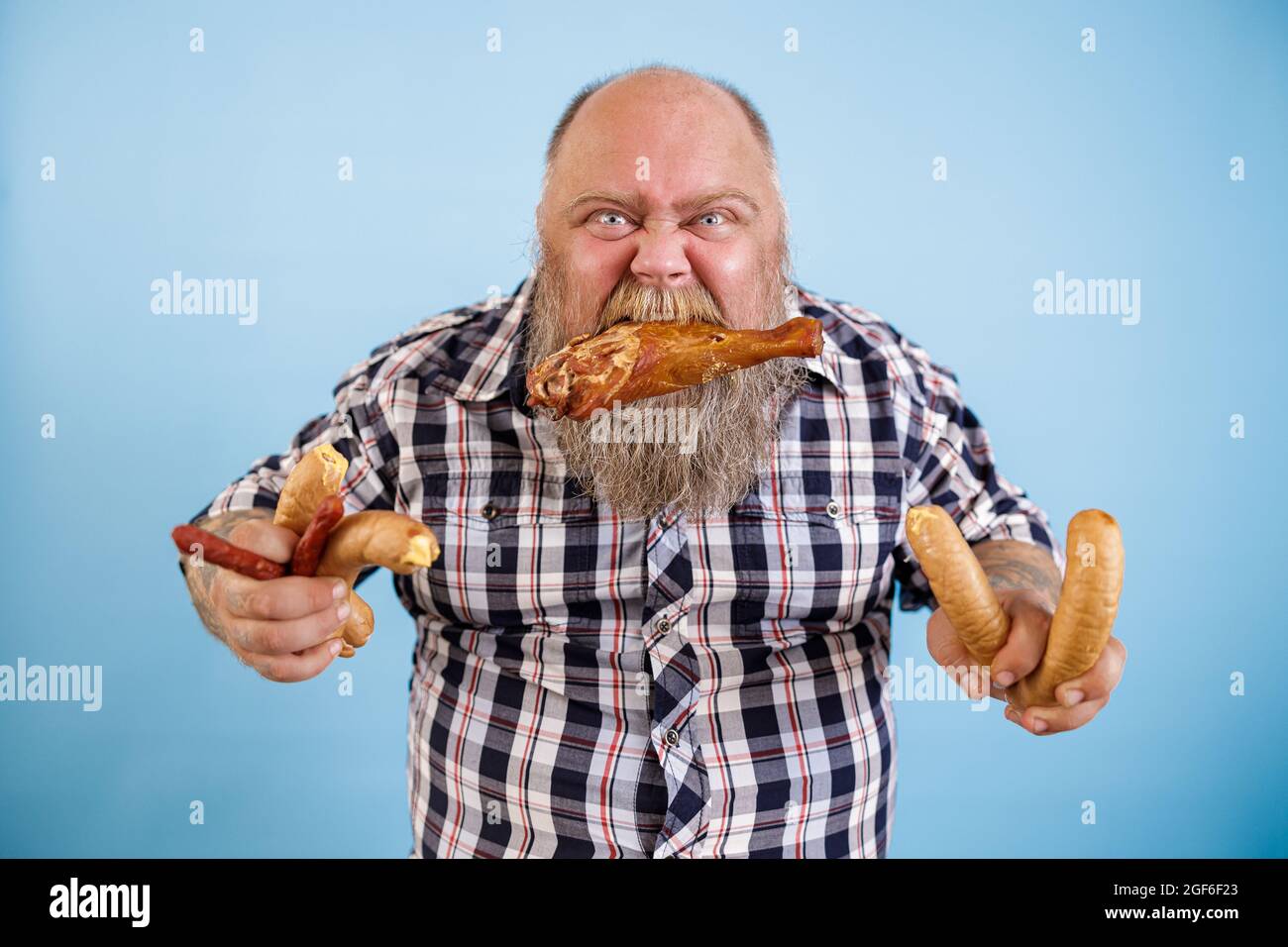 Greedy obese man holds chicken leg in teeth and bunches of sausages on blue background Stock Photo