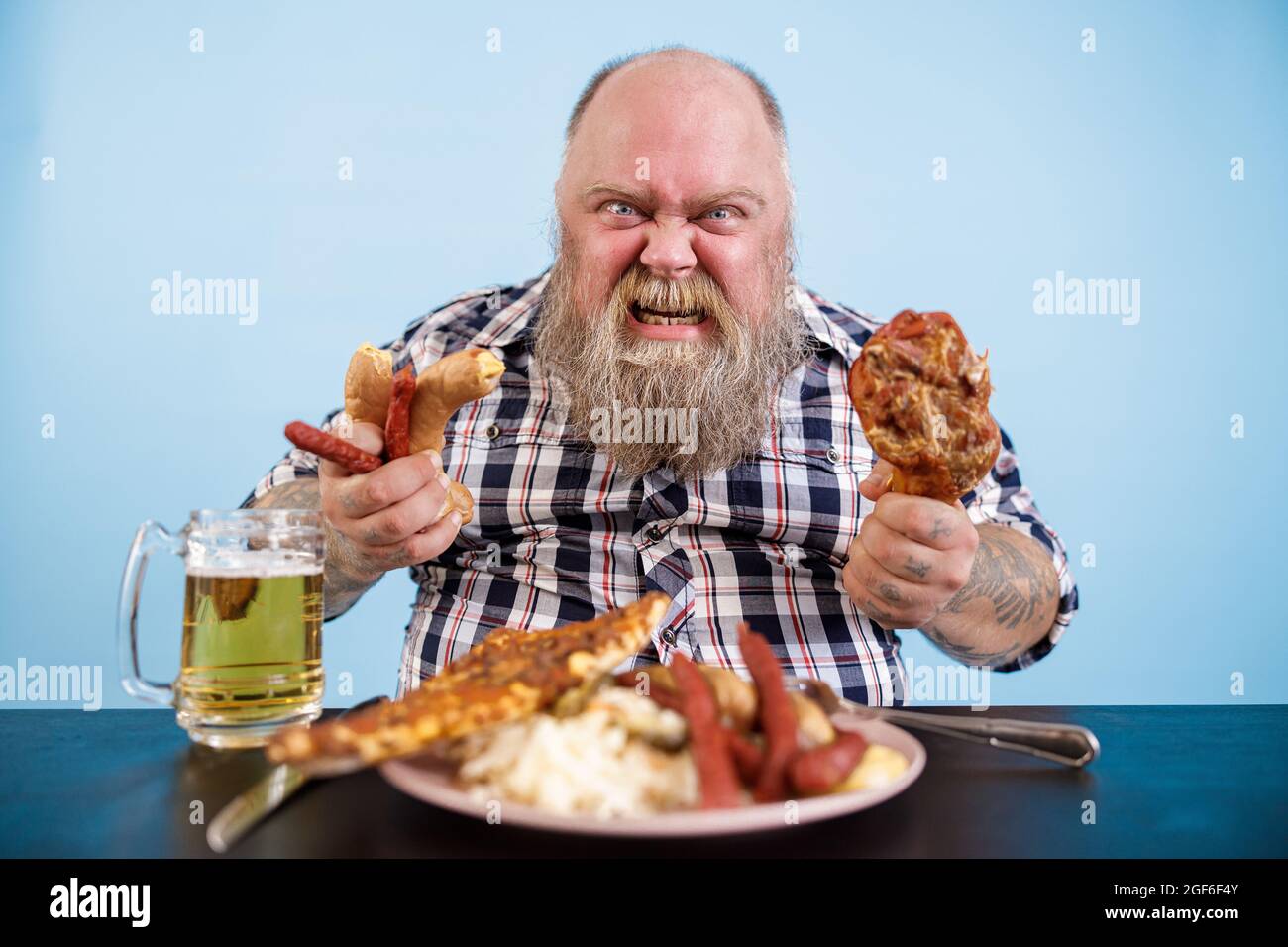 Angry man with overweight holds chicken leg and sausages at table with greasy food Stock Photo