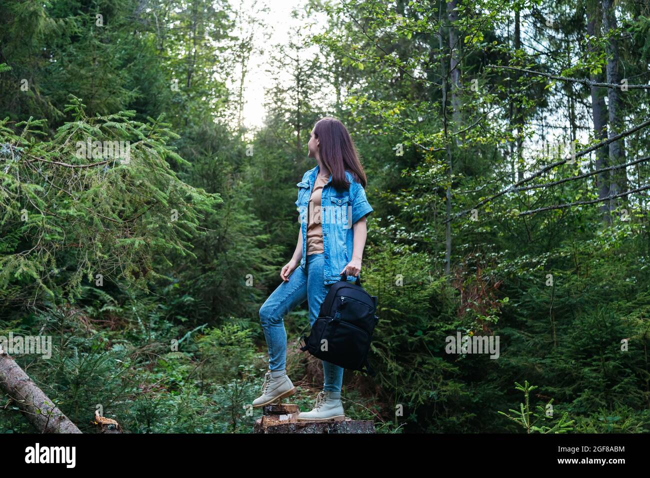 Woman in denim suit in boots with Photo backpack is engaged in hiking in forest. Unrecognizable caucasian young woman walking in nature in park Stock Photo