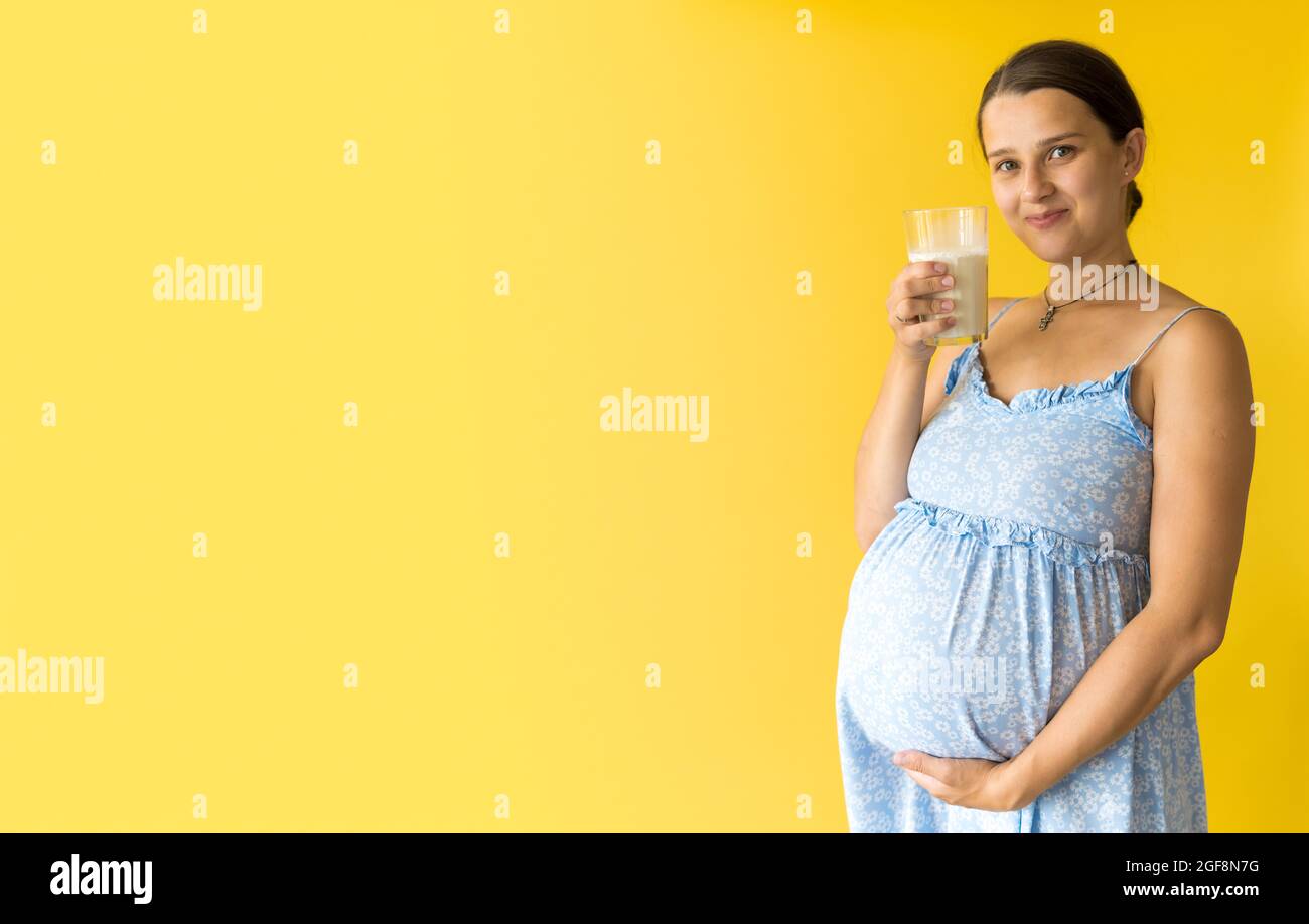 portrait cute beautiful pregnant young pretty woman in floral blue dress holds drink glass of milk rubs tummy on yellow background. Motherhood Stock Photo