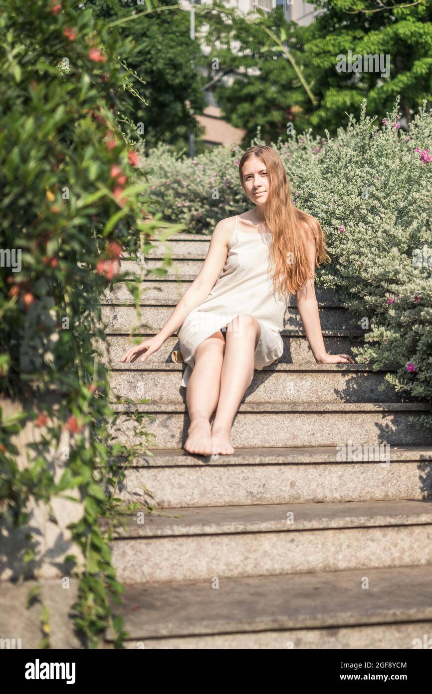 Pretty girl in casual white dress sitting on the stairs in the park. Long blond hair. Romantic summer or spring picture Stock Photo
