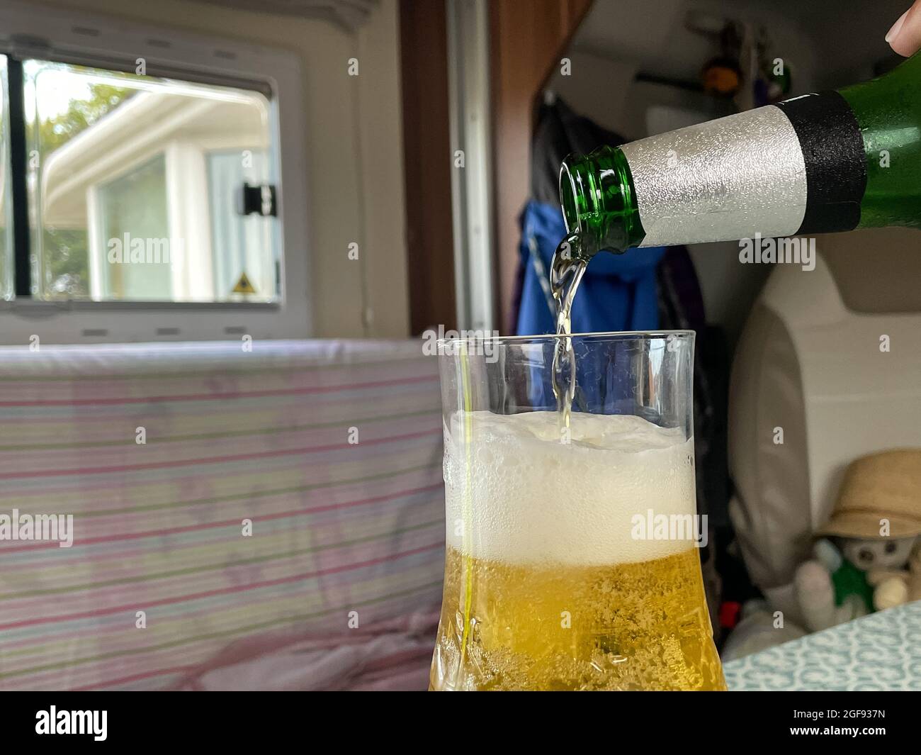 Close view of a green beer bottle with beer pouring into a glass.Lager and frothy head in glass.Interior in a motorhome with window in background Stock Photo