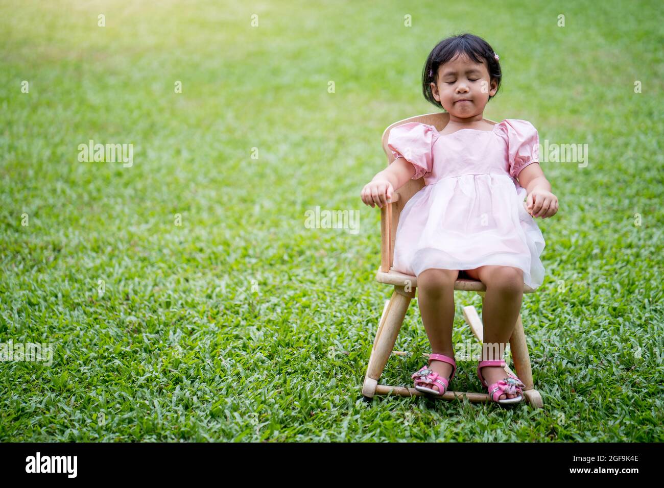 Cute little Asian girl with closed eyes sitting on a chair on the lawn ...