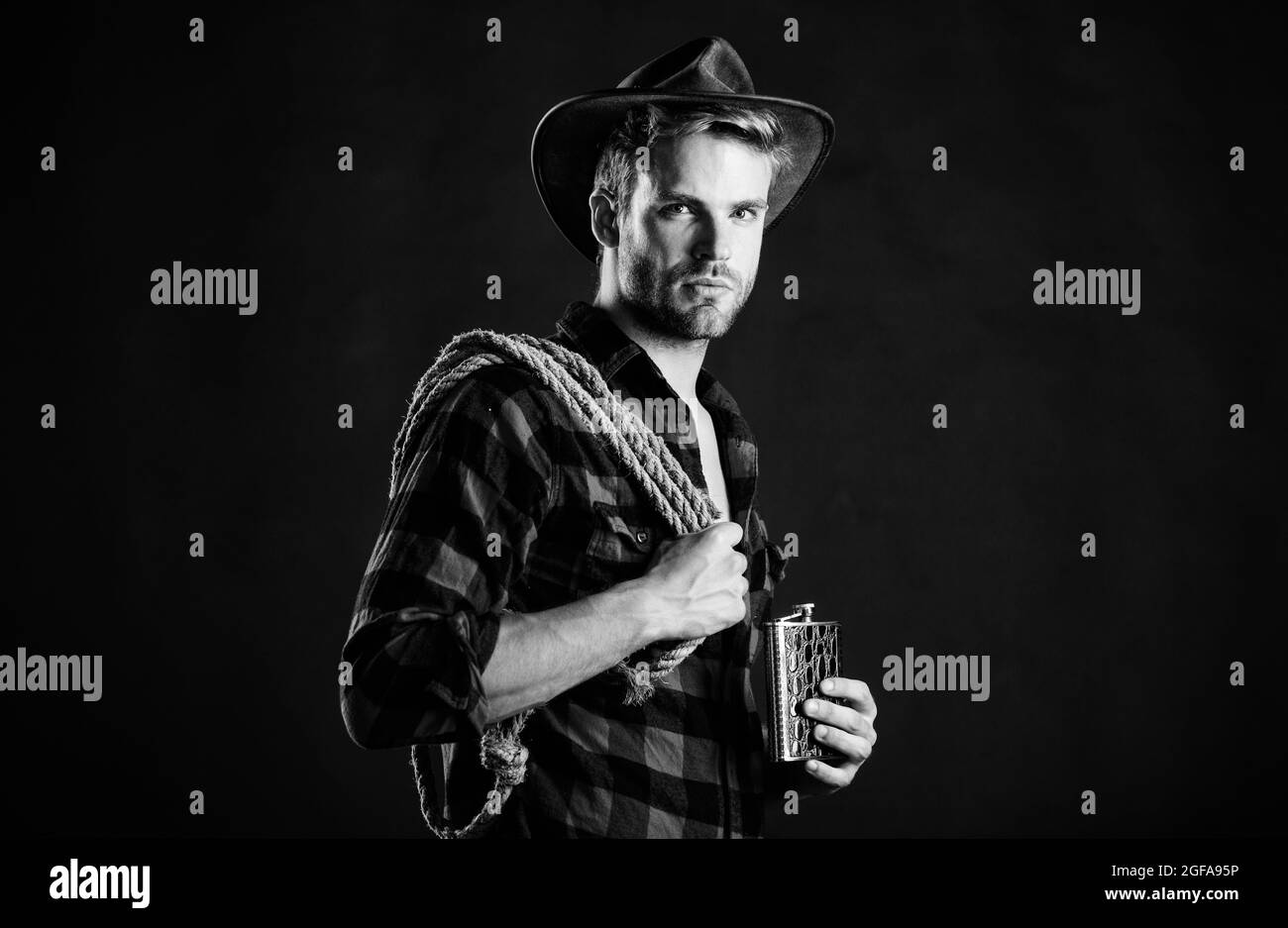 Brutal cowboy drinking alcohol. Western culture. Man handsome unshaven cowboy black background. Western life. Man wearing hat hold rope and flask Stock Photo