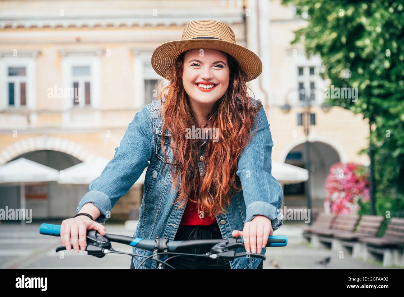 Portrait of smiling long red curly hair caucasian teen girl in a straw hat riding a vintage bicycle in the old town center. Eco transport sharing or c Stock Photo