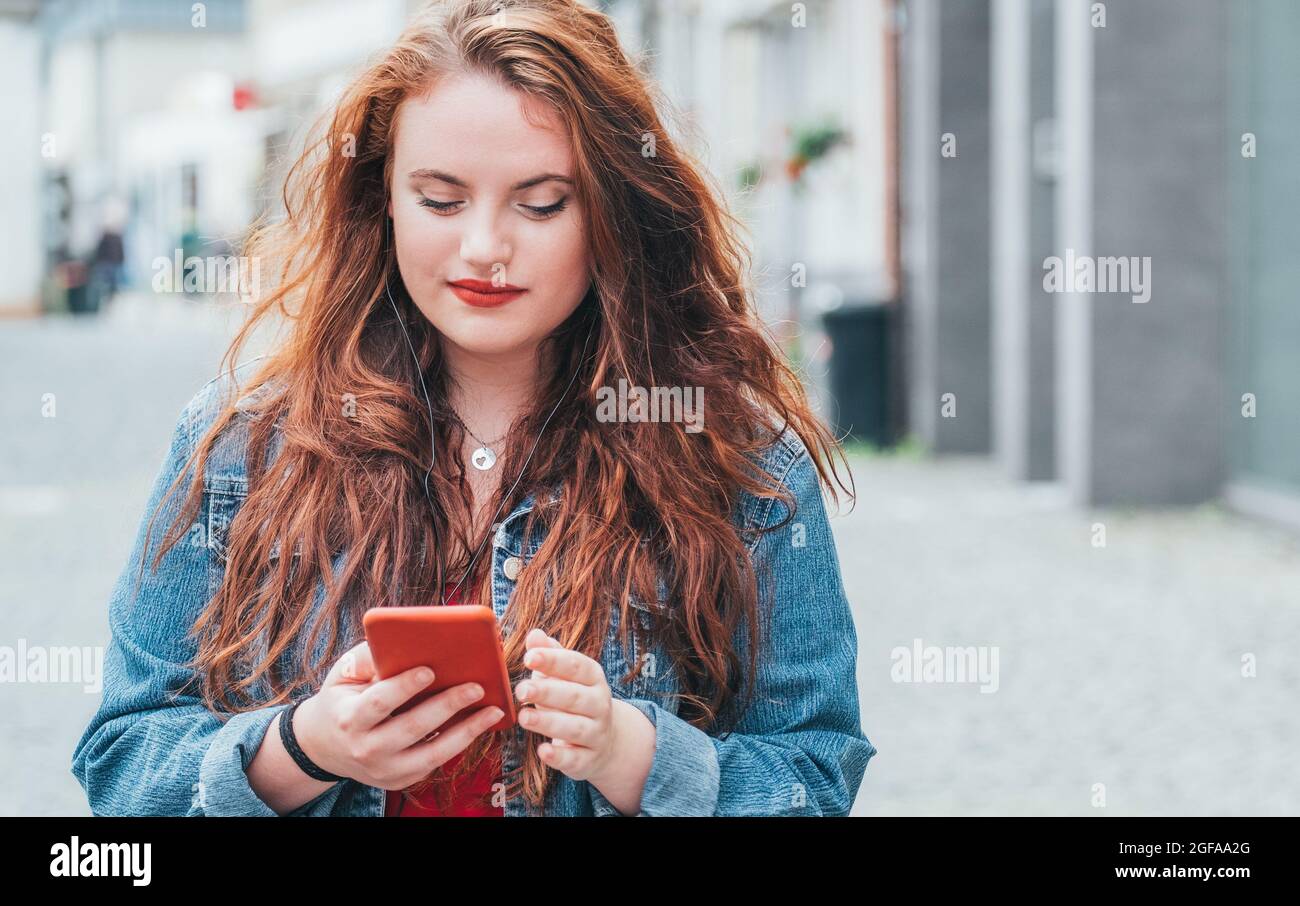 Modern people with technology devices concept image. Portrait of red-curled long hair caucasian teen girl walking on the street and browsing the inter Stock Photo