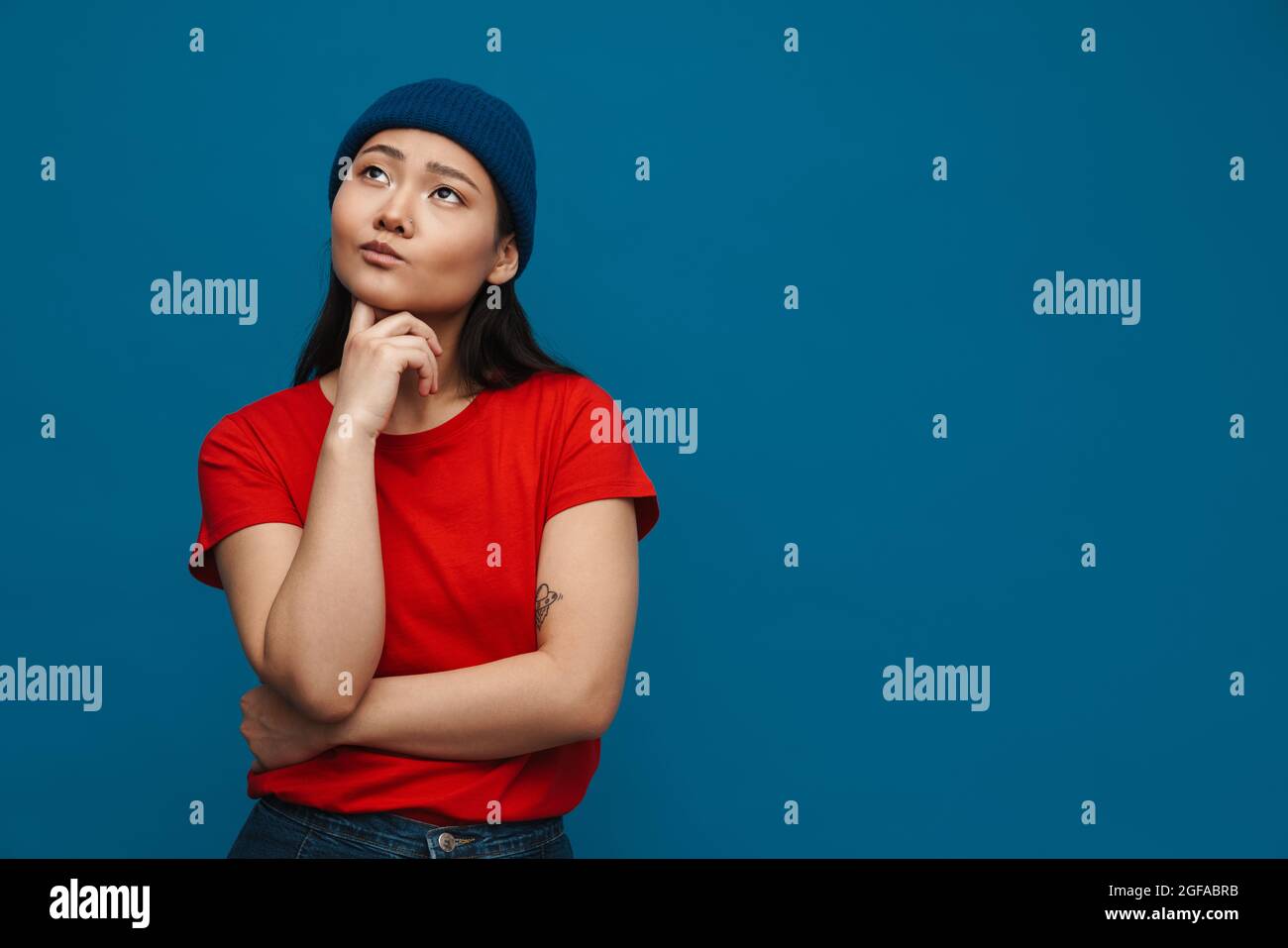 Asian teen girl in hat thinking and looking upward isolated over blue ...