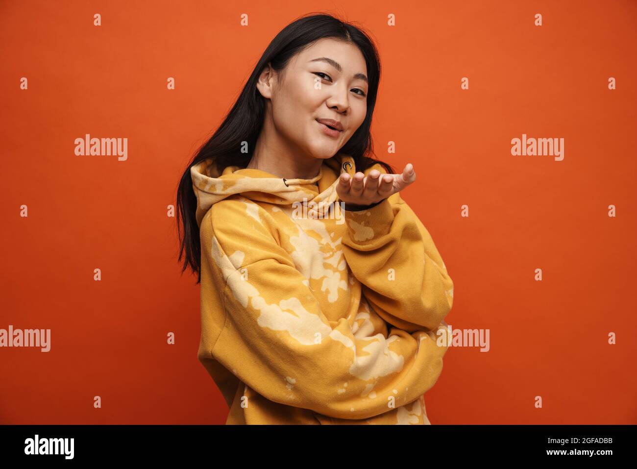 Asian teen girl wearing hoodie blowing air kiss at camera isolated over orange background Stock Photo