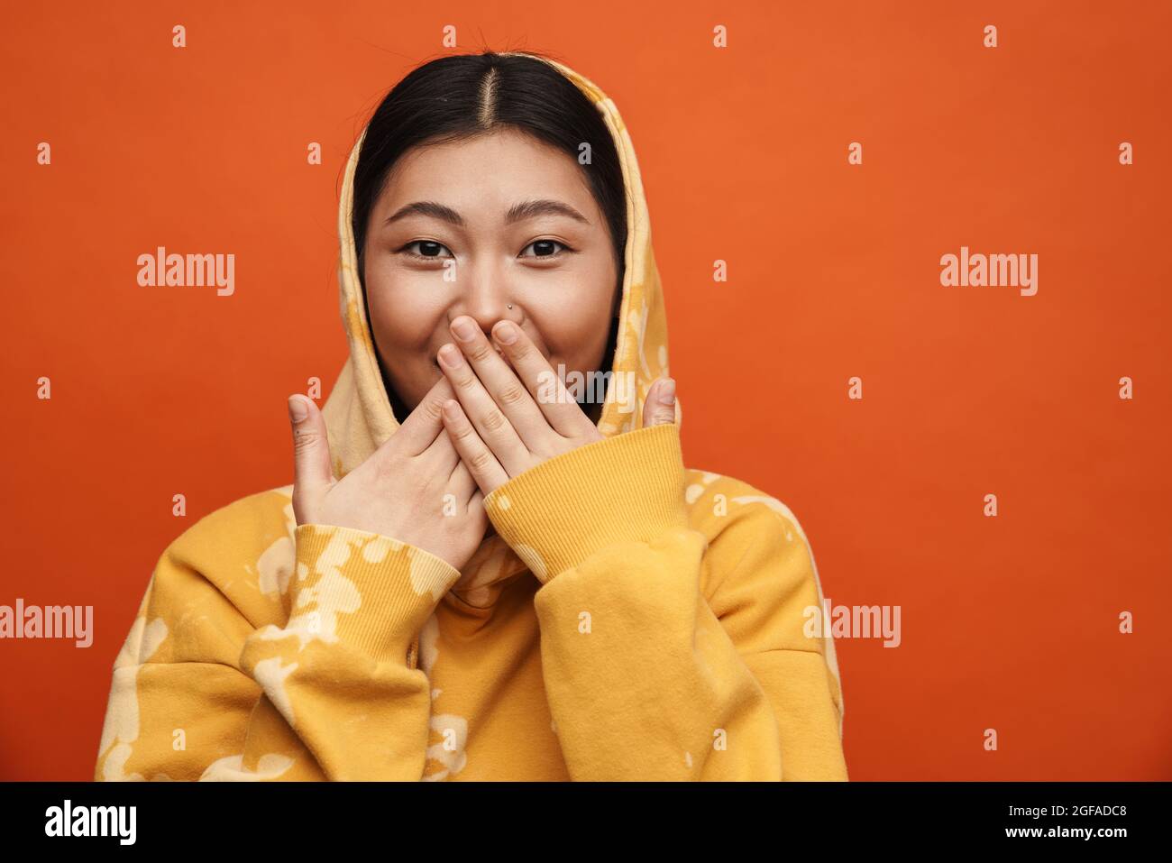 Asian teen girl wearing hoodie covering her mouth isolated over orange background Stock Photo