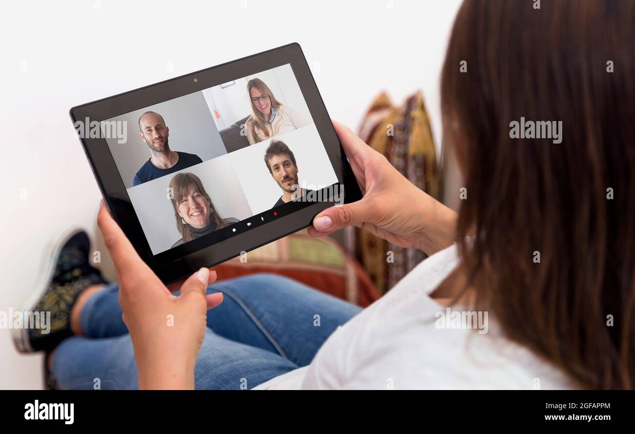 Woman in video conference with a group of people. Girl at home with a tablet in her hands. Young woman resting in a sofa. Millennial girl. Stock Photo