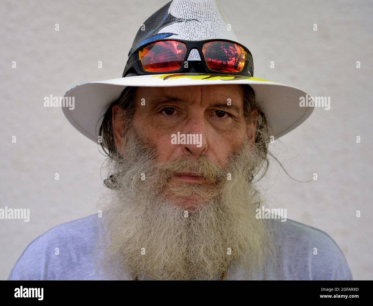 Bearded worried pessimistic elderly Caucasian man wears a Panama hat with colorful sunglasses on the brim and looks at the viewer, white background. Stock Photo