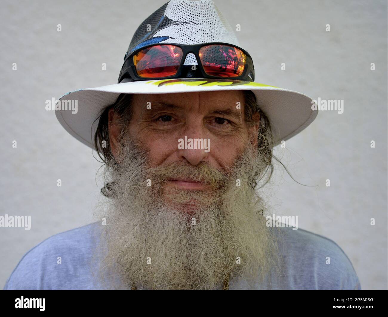 Bearded optimistic elderly Caucasian man grins and wears a painted Panama hat with colorful sunglasses on the brim of his hat, white background. Stock Photo