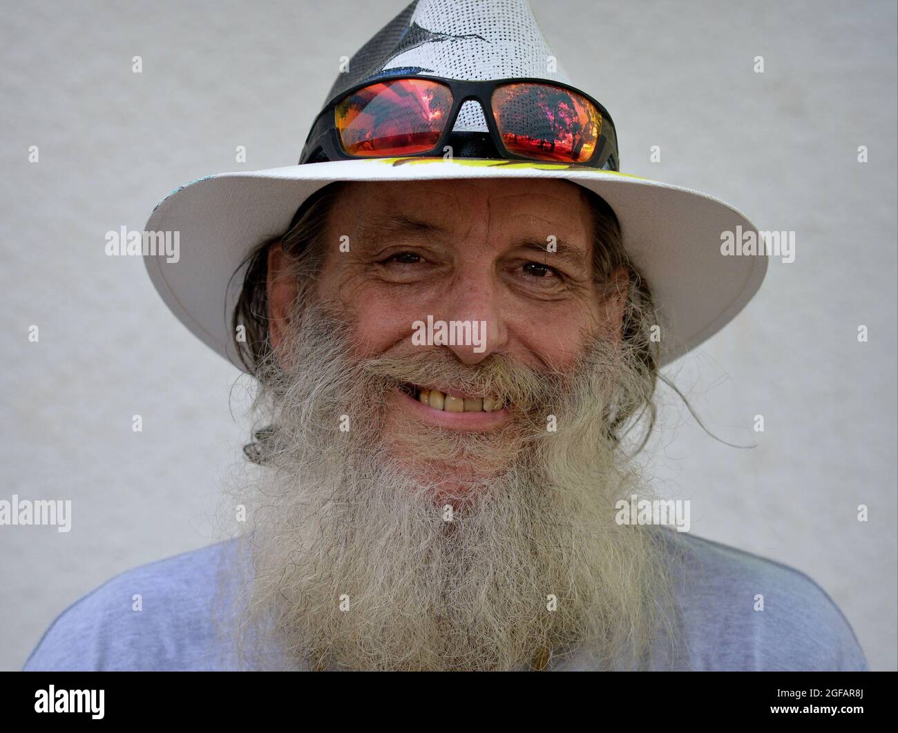 Bearded optimistic elderly Caucasian man smiles and wears a painted Panama hat with colorful sunglasses on the brim of his hat, white background. Stock Photo