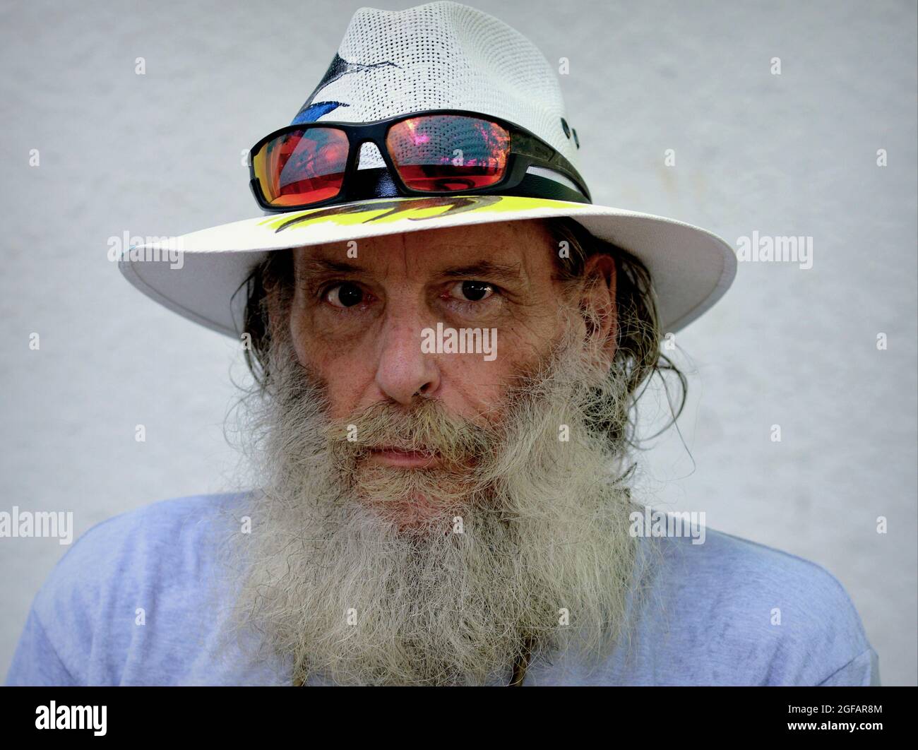 Bearded worried pessimistic elderly Caucasian man wears a Panama hat with colorful sunglasses on the brim and looks at the viewer, white background. Stock Photo