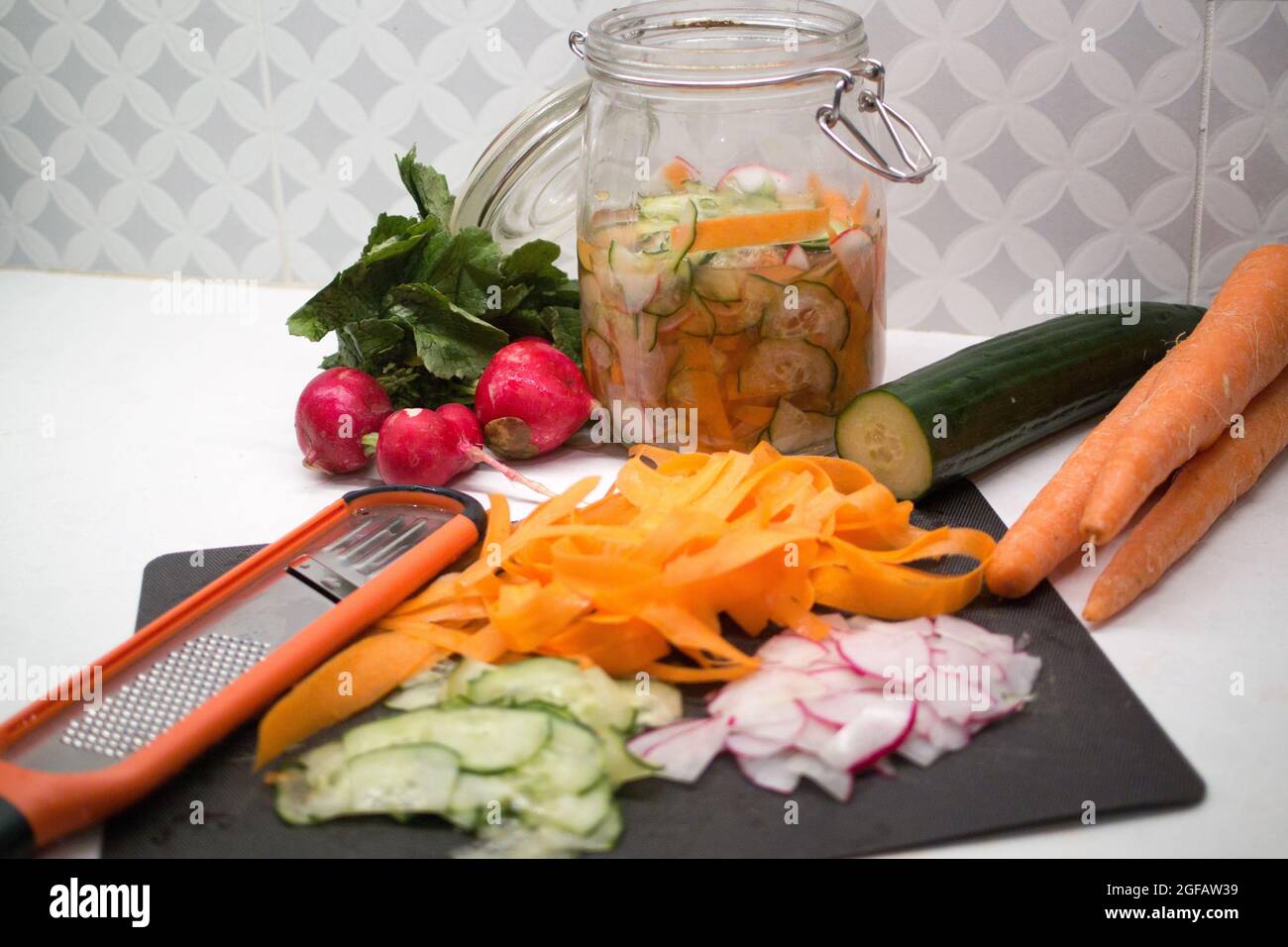 Carrots, cucumbers and radishes being thinly sliced by microplane to preserve in large kilner jar with pickling vinegar. Stock Photo