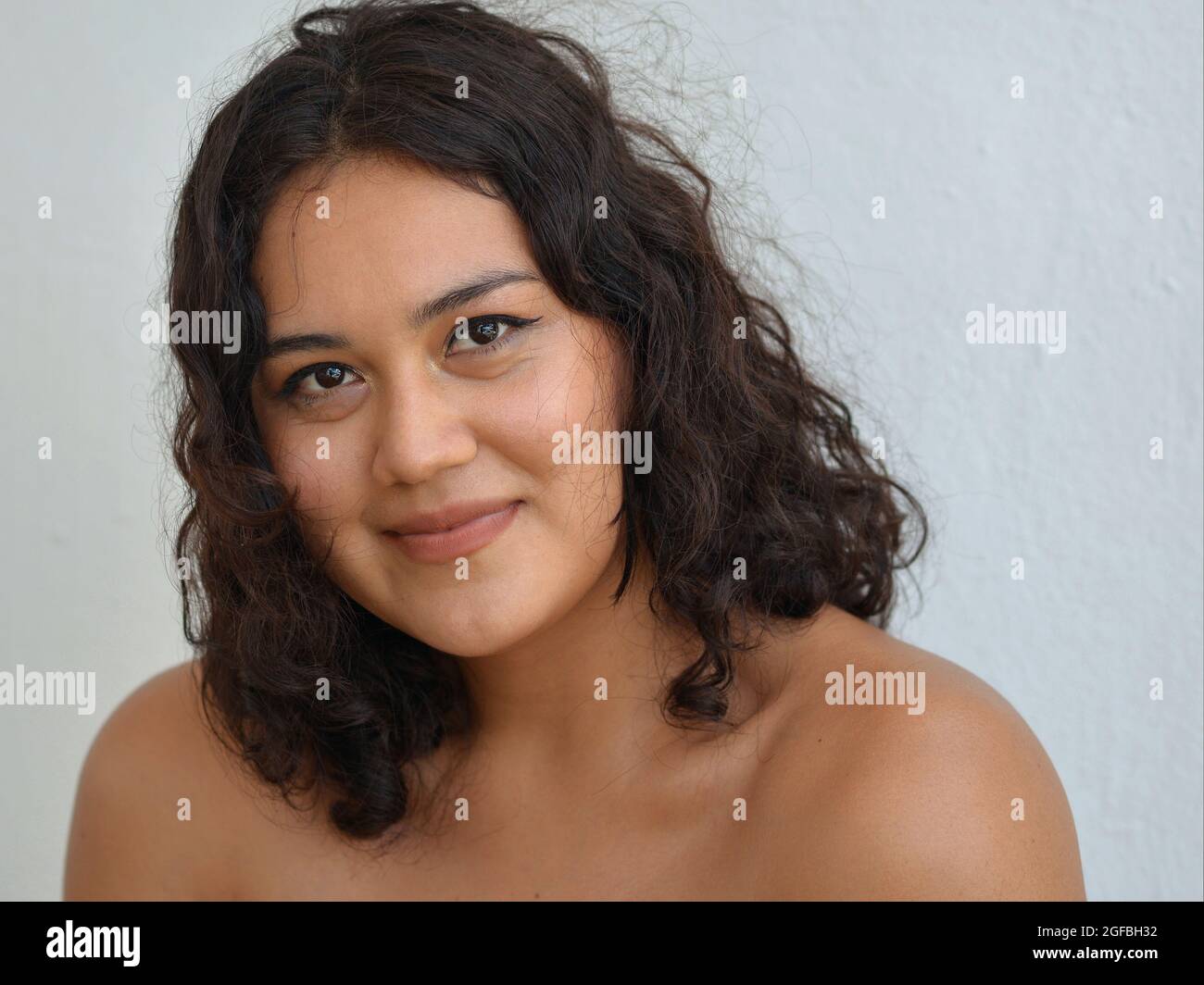 Young beautiful Mexican Latina woman with curly long hair, smiling brown eyes and bare shoulders poses for the camera in front of a white background. Stock Photo