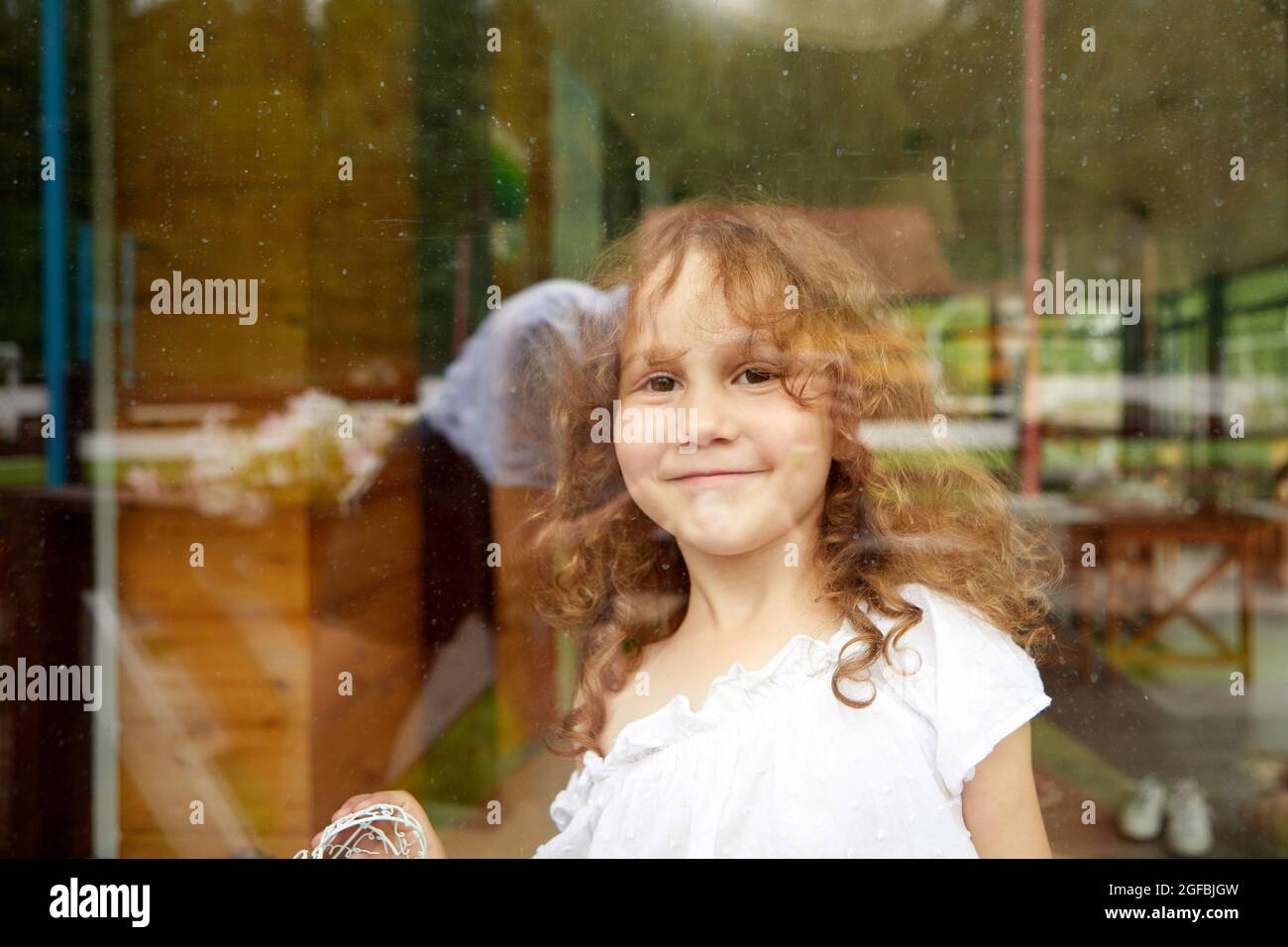 Cute happy girl with curly hair smiling and looking at camera while standing behind transparent glass Stock Photo