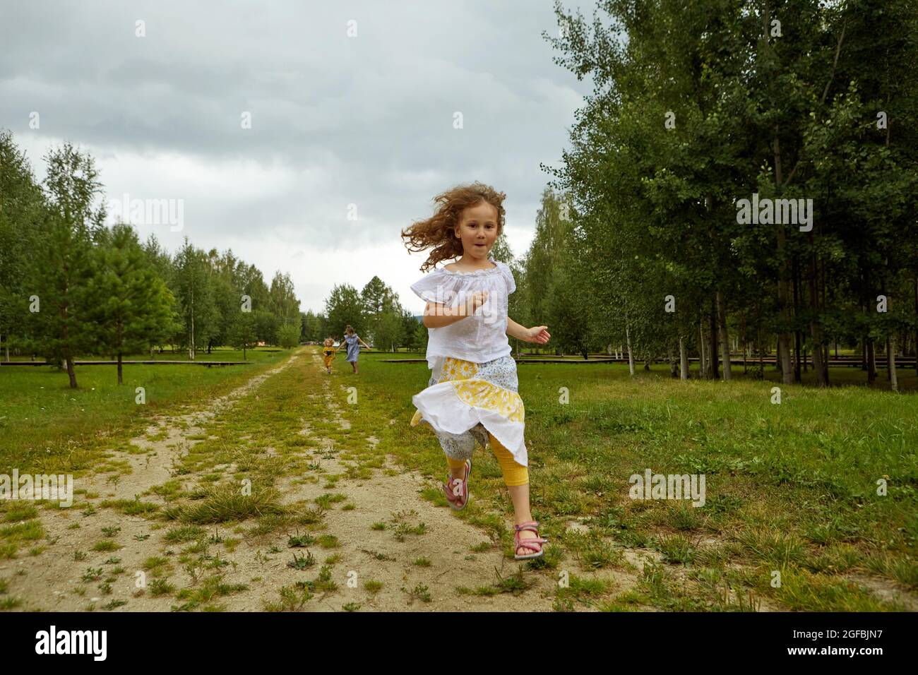 Funny girl in skirt and blouse running on road towards camera against cloudy sky while spending weekend day near grove in nature Stock Photo