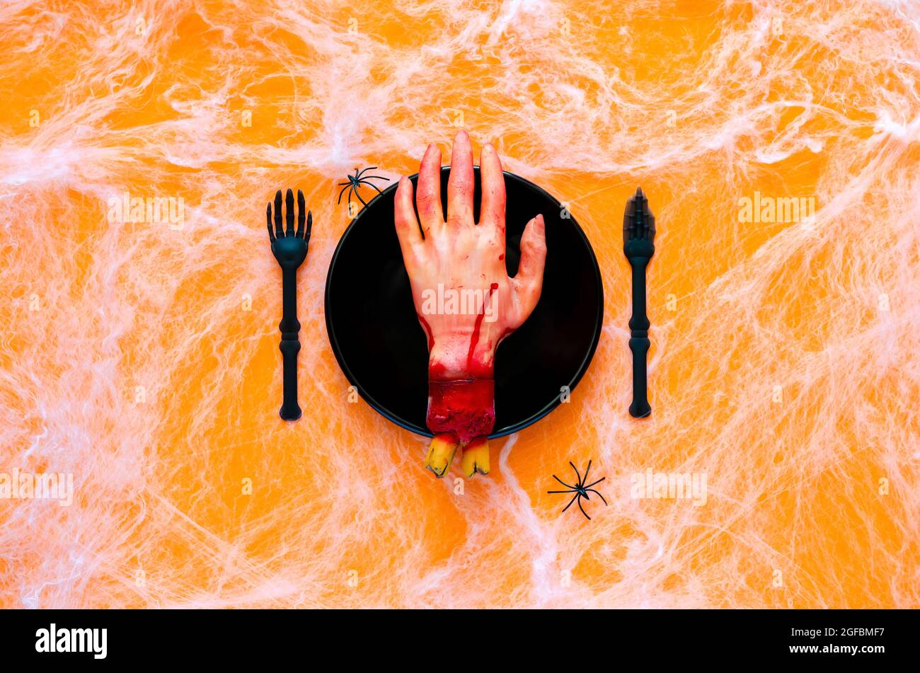 Scary halloween dinner party concept with fake cutting hand on black plate, knife and fork with spider and cobweb on orange background. Stock Photo