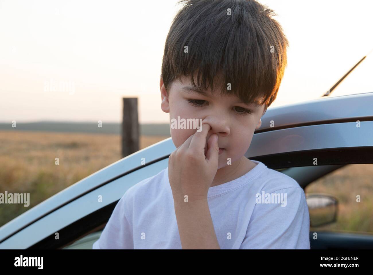 Boy picking his nose next to a car at sunset. Dry and hot weather brings malaise and respiratory problems. Stock Photo