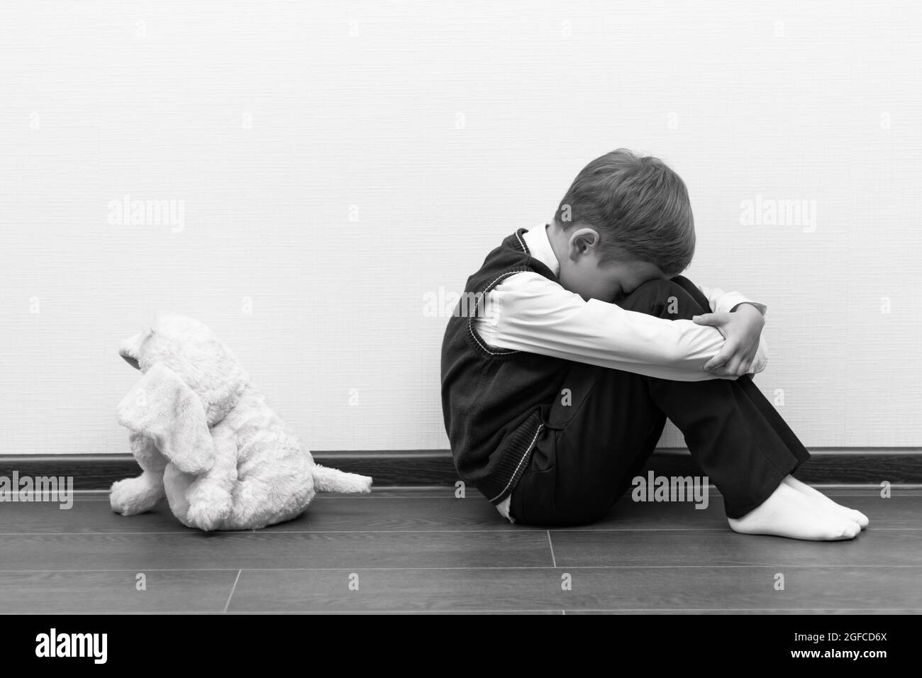 A little boy of six is offended and sits on the floor against a white wall next to a soft toy with his hands closed. Selective focus. Close-up Stock Photo