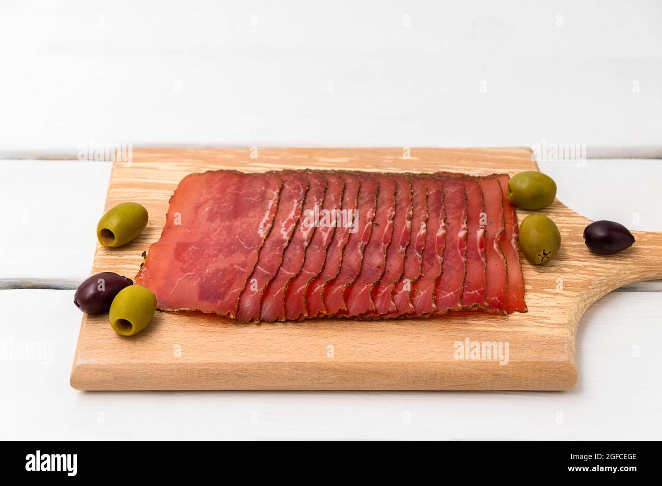 Fin slices of dried meat on cutting board with olives on white wooden table Stock Photo
