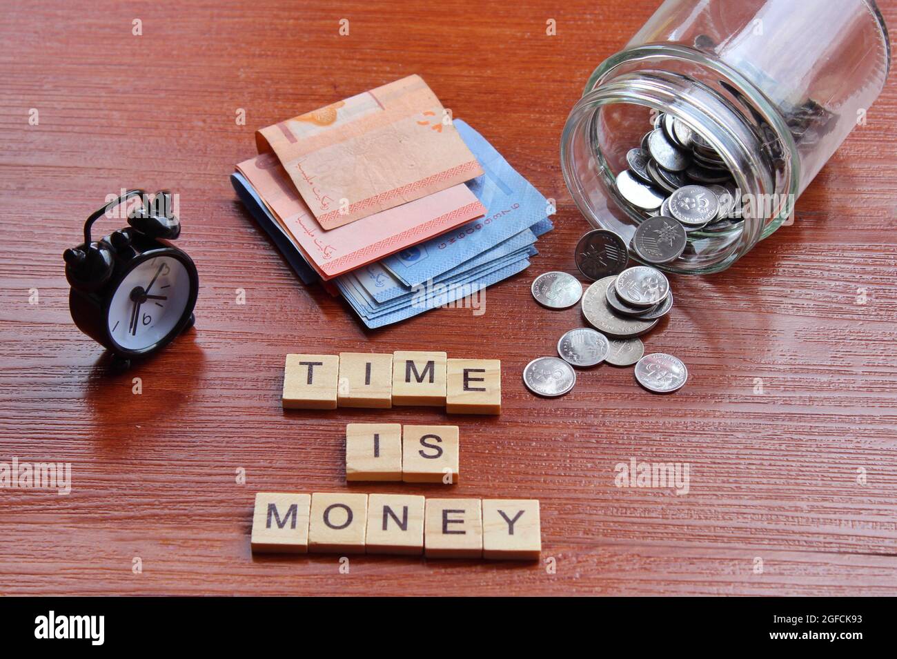 Close up of alarm clock, coins, banknotes and wooden cube with text TIME IS MONEY Stock Photo