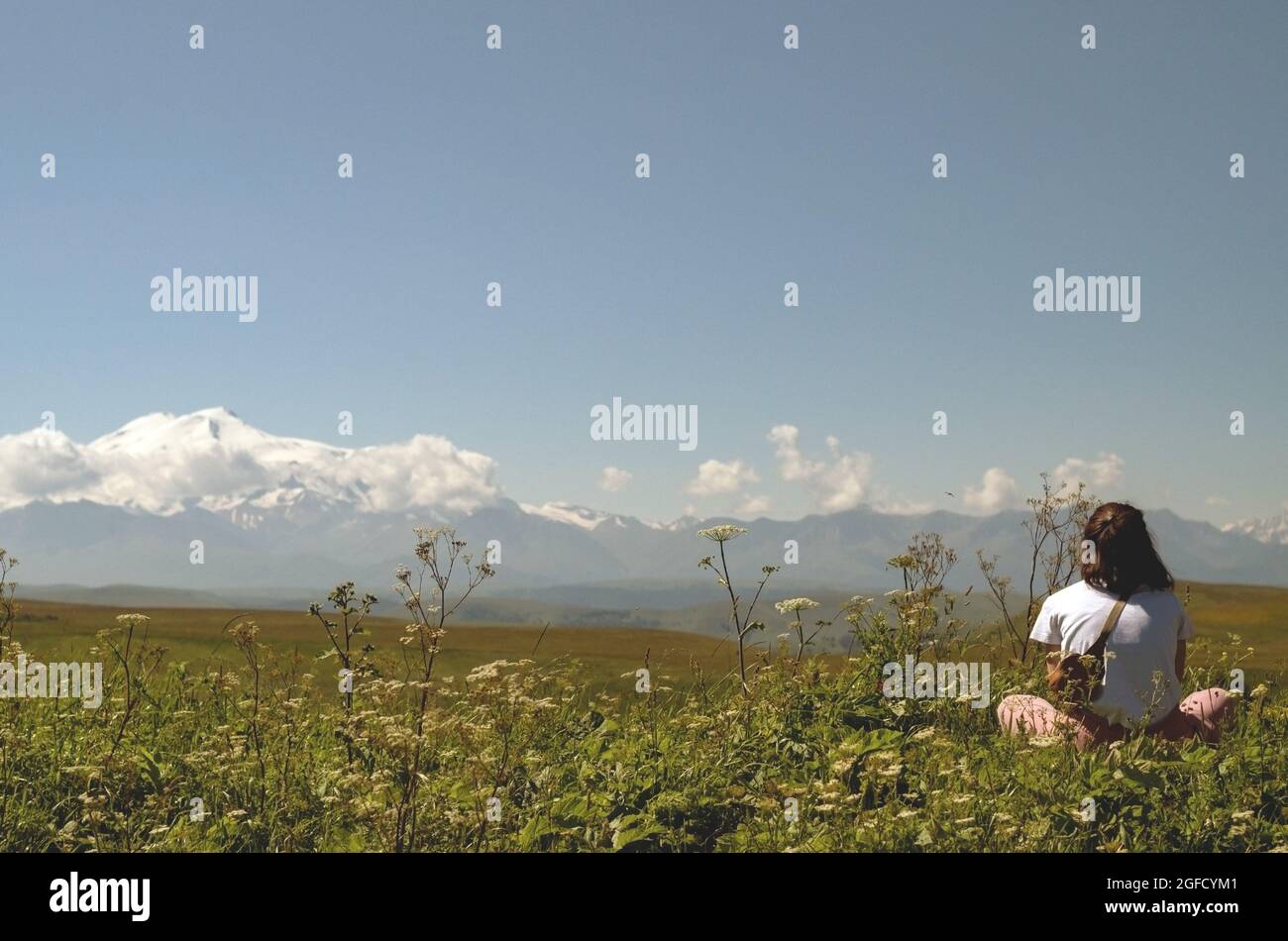 A girl sits in a meadow and looks at a beautiful mountain landscape.Mountain travel concept Stock Photo