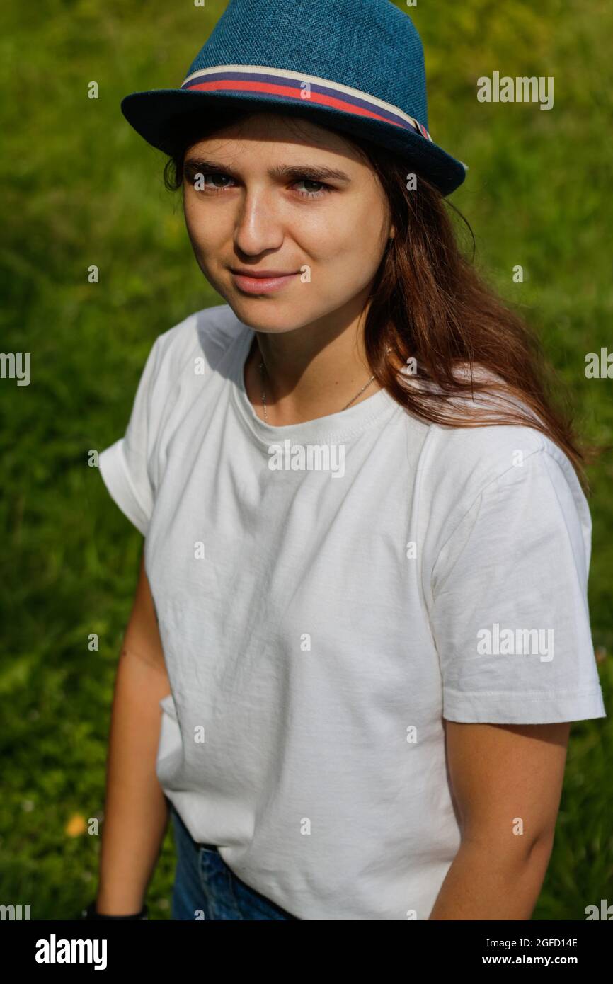 Defocus close-up portrait of a tired young woman with brown hair wearing a hat outdoors. Green nature background. Looking at camera. Vertical. Out of Stock Photo