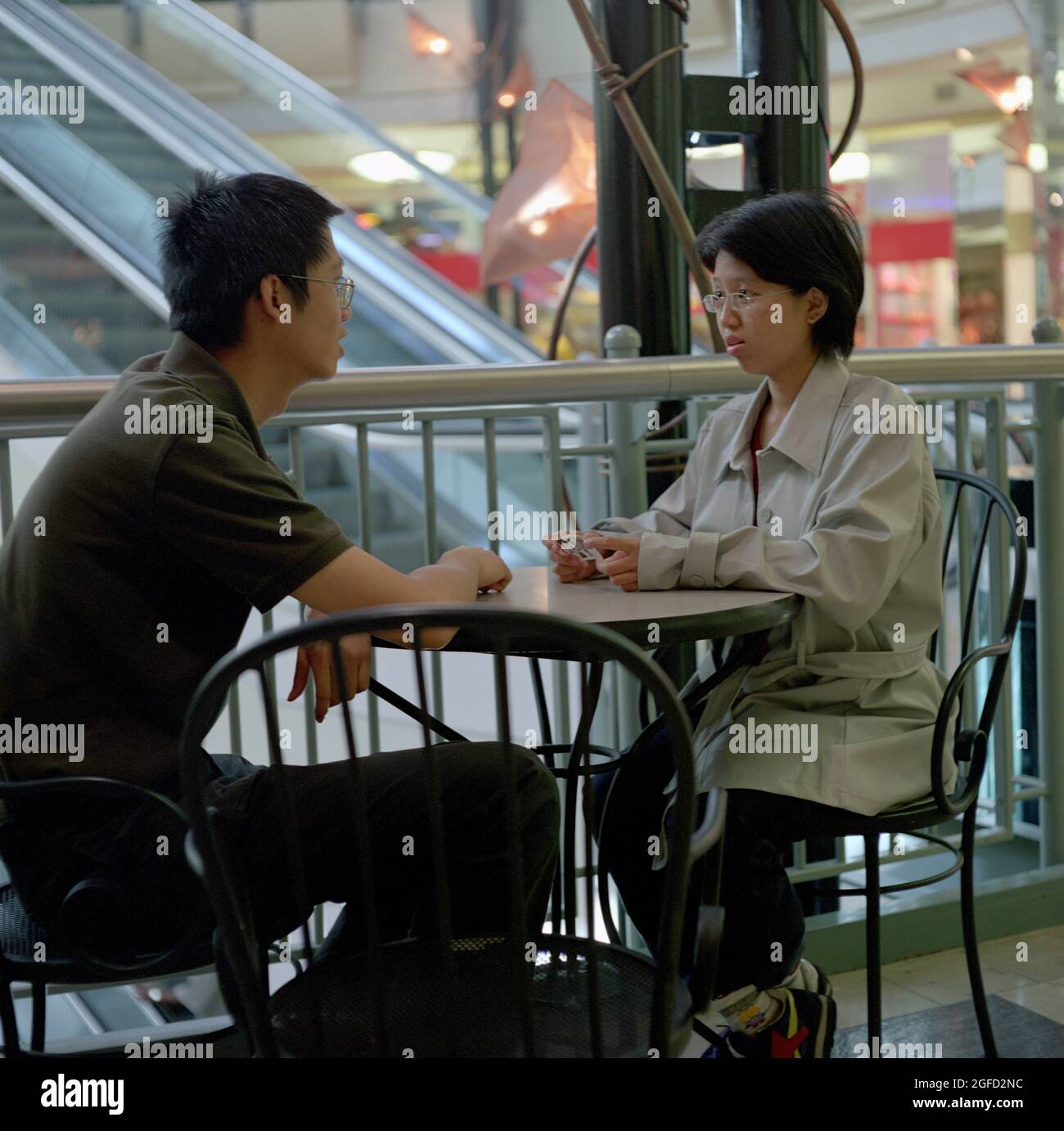 An asian American couple discuss world events at a small table near the escalator in the mall. Stock Photo
