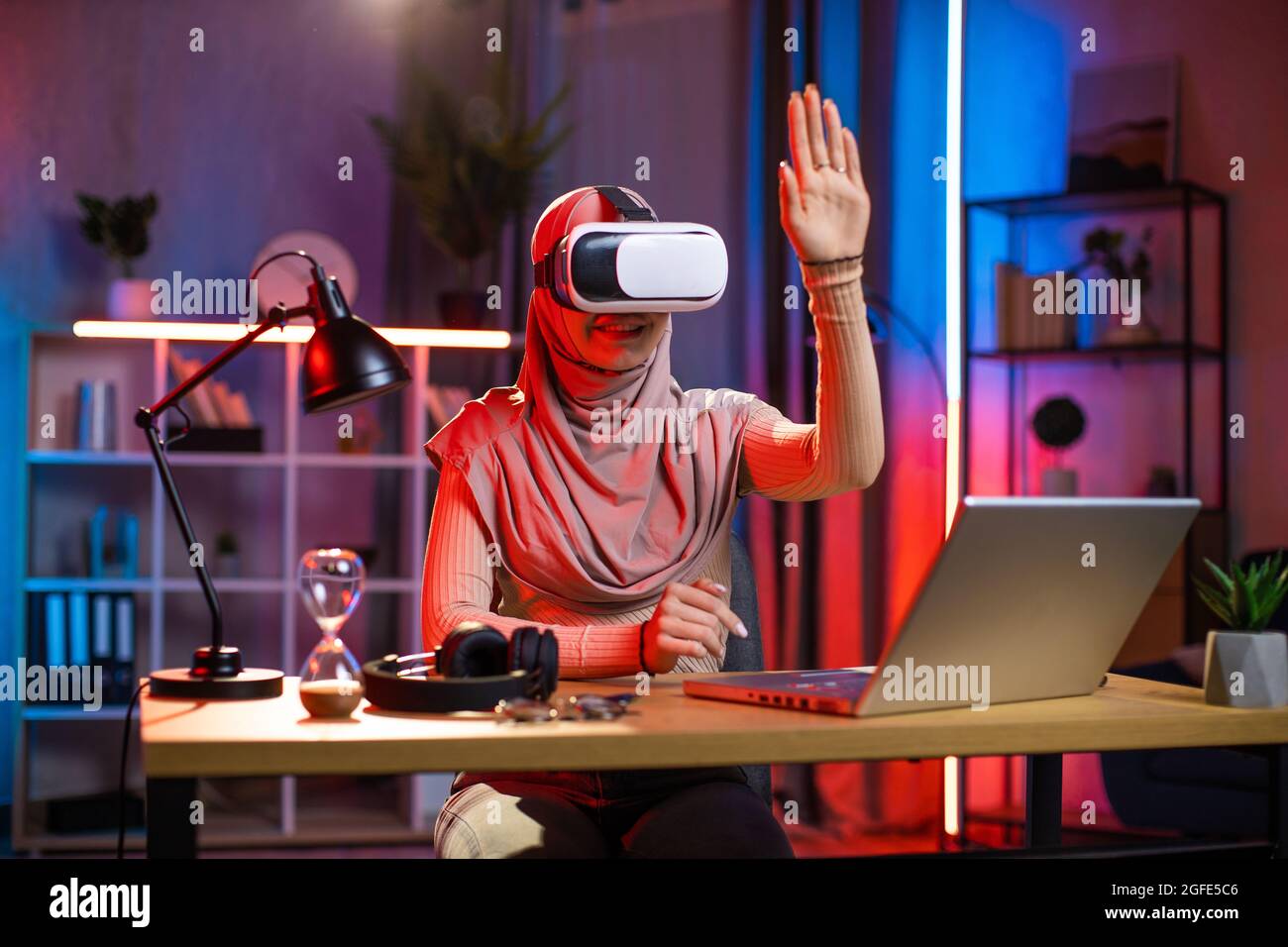 Young arabian woman sitting at home office and using VR headset and laptop for work during evening time. Concept of people, technologies and freelance Stock Photo