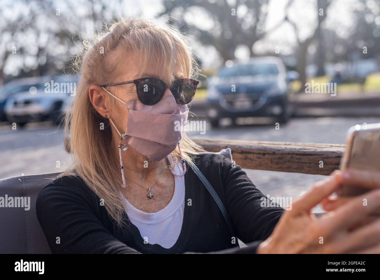 Mature adult woman smiling with glasses wearing face mask looking at smartphone. Stock Photo