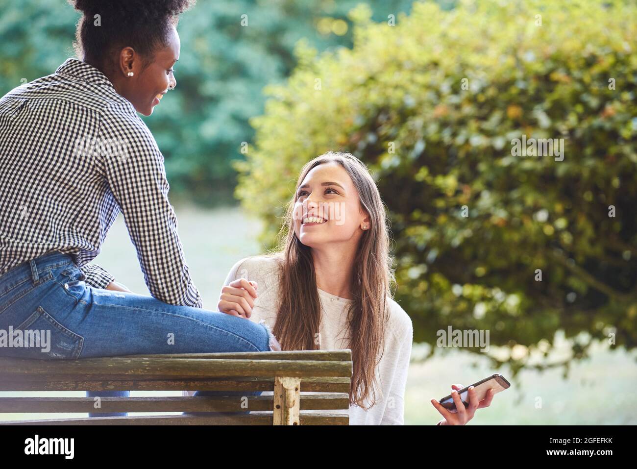 Smiling young friends looking at each other while sitting on bench Stock Photo