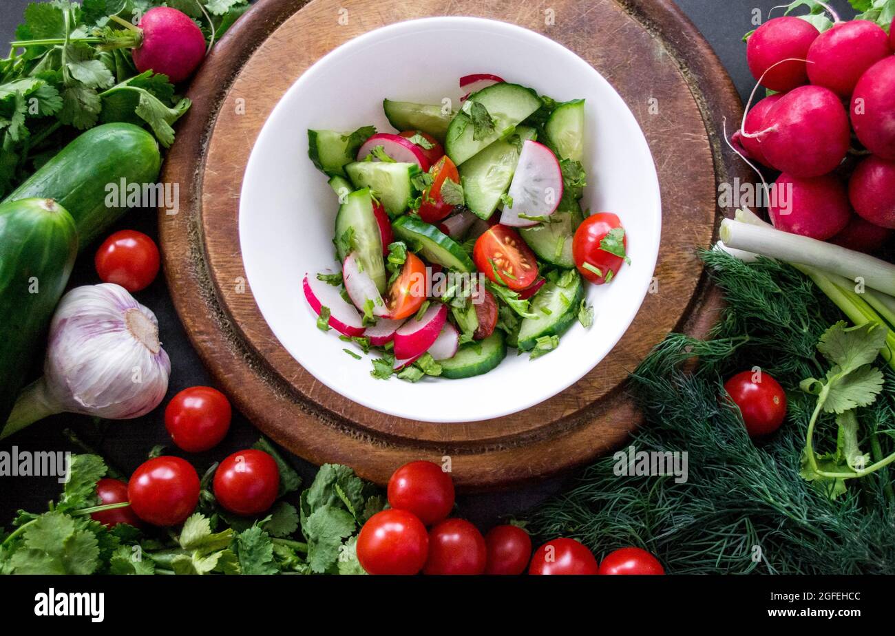 A pile of fresh, ripe vegetables: radishes, tomatoes, cucumbers, herbs, and garlic are arranged around a round wooden chopping board. White bowl veget Stock Photo