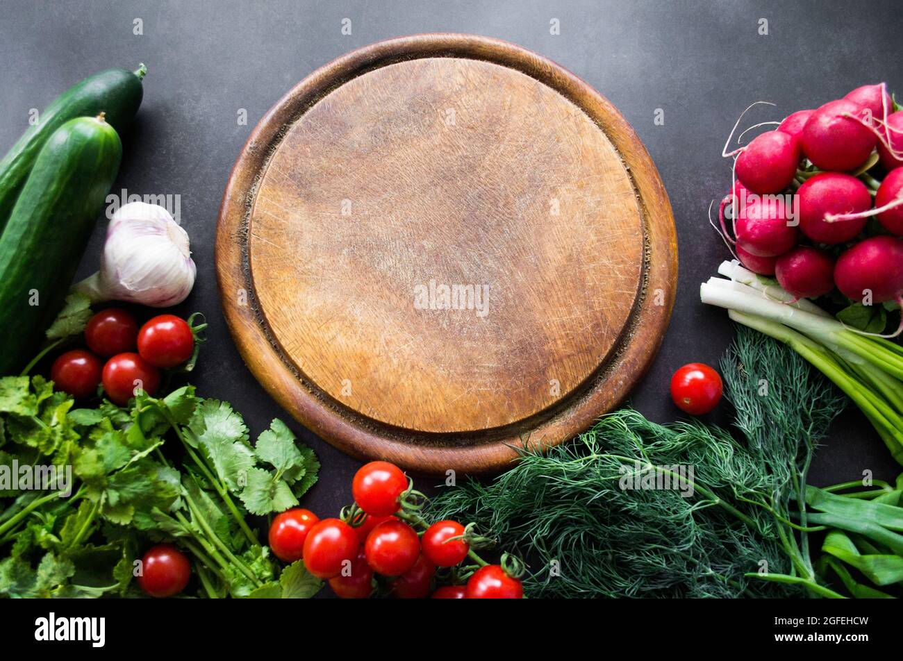 Top view a pile of fresh, ripe vegetables: radishes, tomatoes, cucumbers, herbs, and garlic are arranged around a round wooden chopping board. Copy sp Stock Photo