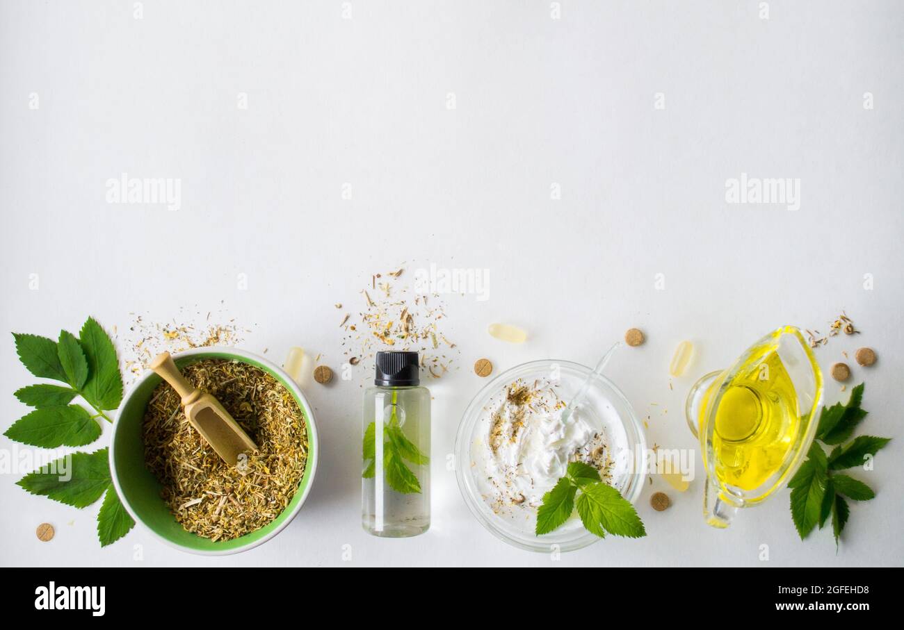 Alternative medicine, herbal extract: white background, jar of oil over a bowl with white essence. Bowl with ground dry medicinal herbs, green leaves Stock Photo