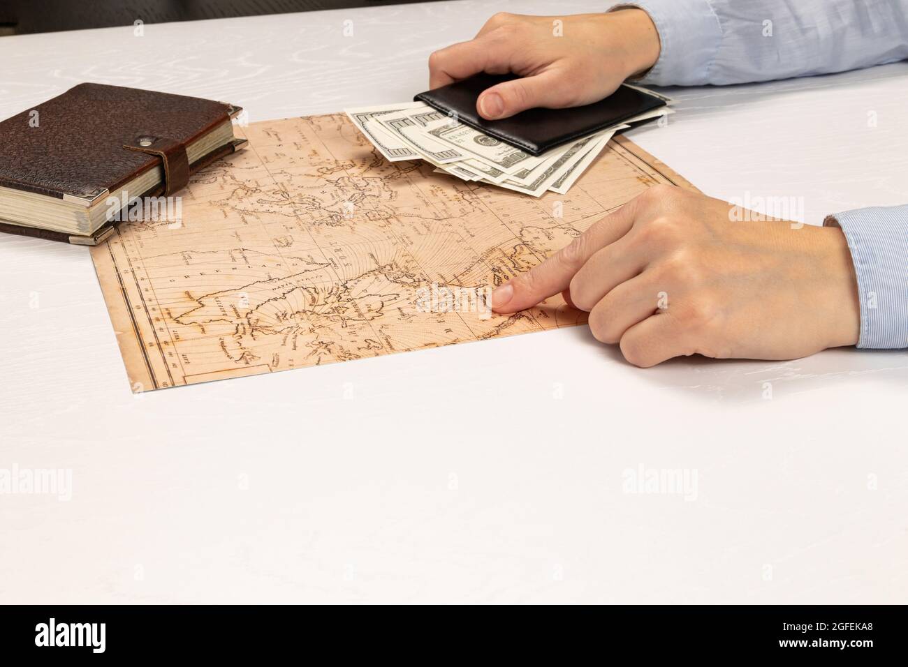 female hands hold a black leather wallet with money and show the resting place on the world map. brown diary on a white wooden table Stock Photo