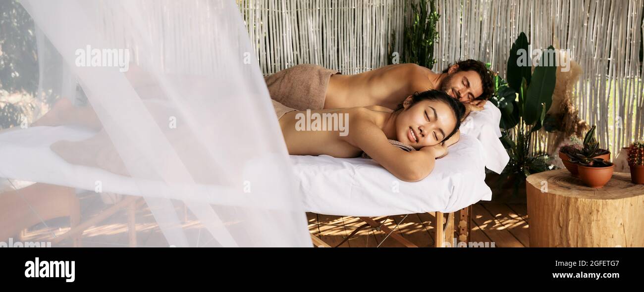 Multi-ethnic couple having relax during back massage lying with eyes closed on massage tables at romantic spa resort, summer outdoor. Side view Stock Photo